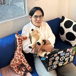 รูปภาพของ Thanakorn Klumpoo ใส่แว่นกลมข้างเหลี่ยมข้าง มี Laptop อยู่ที่หน้าตัก นั่งในคาเฟ่และถือตุ๊กตา